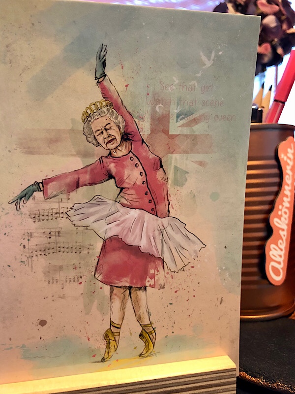 Postkarte von der Queen im Tutu mit geschlossenen Augen, auf Spitze tanzend, Zeichnung, rosa Kleid, im HIntergrund Stifthalter mit Button "Alleskönnerin"